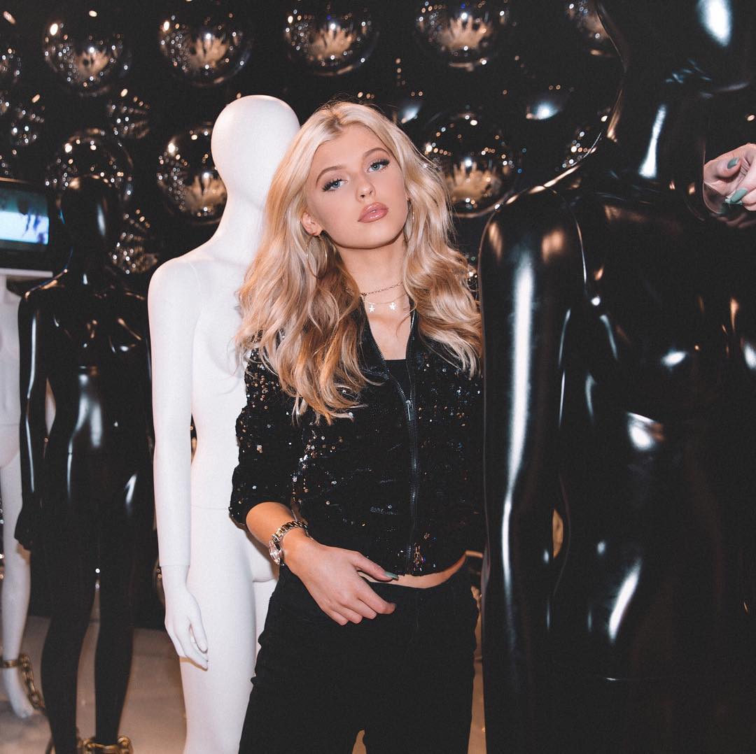 a woman with blonde hair stands between a white mannequin and a glossy black mannequin. she wears a sparkly black jacket, black pants, and jewelry. the background features disco balls.