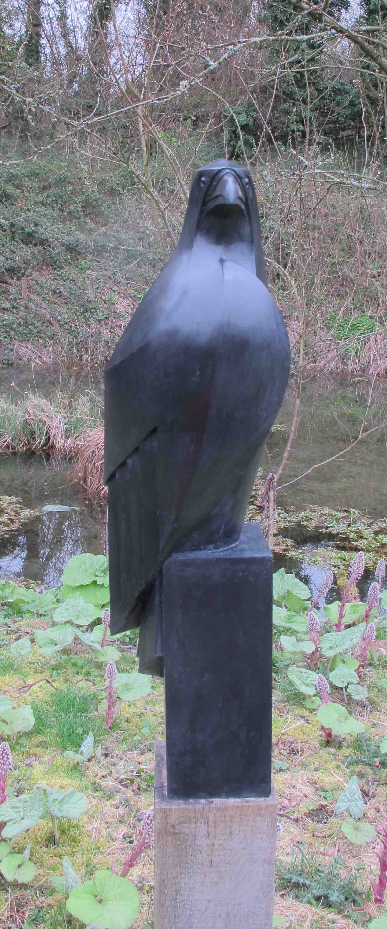 a tall, modern sculpture of a bird, possibly a crow, stands in a gardenlike setting. the sculpture is dark in color and appears to be made of stone or metal with geometric shapes. it is placed on a square pedestal and sits among lush plants, with a pond in the background. the scene suggests a peaceful, natural environment.