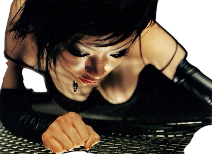 a closeup shot of a young woman with dark hair and intense eyes. she is wearing black clothing and gloves and is leaning forward, her gaze directed downward. the background is a metallic grid. the image is high contrast with a moody and dramatic atmosphere. the focus is sharp on the woman's face and hands.