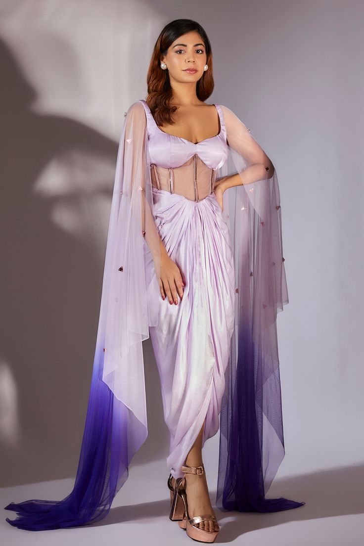 the image showcases a female model elegantly dressed in a lavender satin dhoti saree. the saree features a fitted corset bodice with sheer detailing, creating a unique and modern silhouette. a matching, ombre net dupatta (shawl) in shades of lavender and purple is draped around the model's shoulders and trails on the floor. the dupatta has subtle embellishments. the model wears rose gold high heels and has her hair styled in soft waves.  the backdrop is a simple, lightgrey studio setting, allowing the outfit to be the focal point. the overall aesthetic is stylish and sophisticated, suitable for a formal occasion or celebration.
