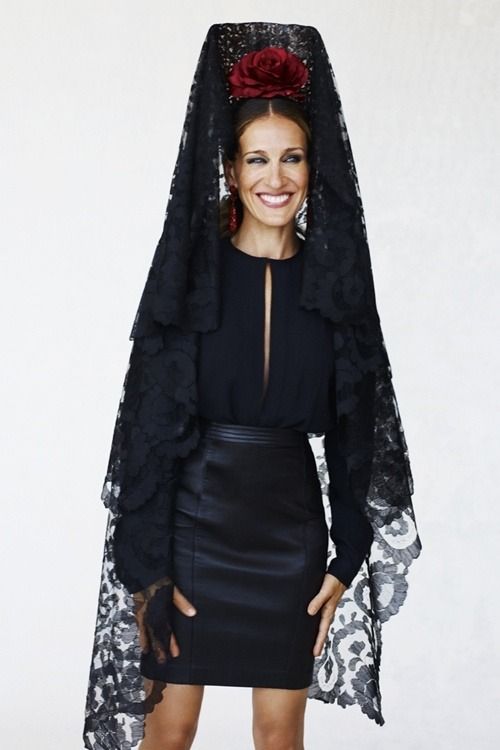 a woman stands against a white background, smiling and looking directly at the camera. she is wearing a black dress with a keyhole neckline and a black leather pencil skirt. a black lace mantilla with a large red rose adorns her head. she is also wearing red earrings.