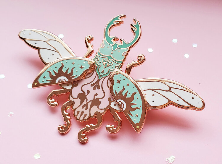 the image is a closeup shot of an enamel pin depicting a fantastical beetle. the pin is set against a light pink background with a few scattered glitter pieces. the beetle is highly stylized with intricate designs and a combination of colors including rose gold for the metal outlines and details, mint green, white, and pink for the enamel filling. it has large, decorative horns, patterned wings with eye symbols and star details, and stylized legs. the overall aesthetic is whimsical and artistic.