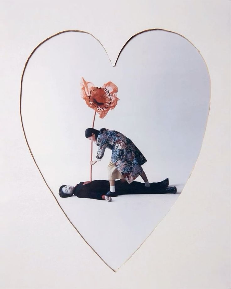 the image is a photograph framed within a heartshaped cutout on a white background. the photo itself depicts a surreal scene. a woman in a patterned coat is bending over a man lying on the floor, apparently "stabbing" him with a long, slender stem of a large, stylized red poppy.  the overall style is minimalist and evocative, suggesting a symbolic narrative about life, death, relationships, or possibly a commentary on fashion and performance art. the muted color palette and stark setting contribute to the image's dramatic and somewhat unsettling mood.
