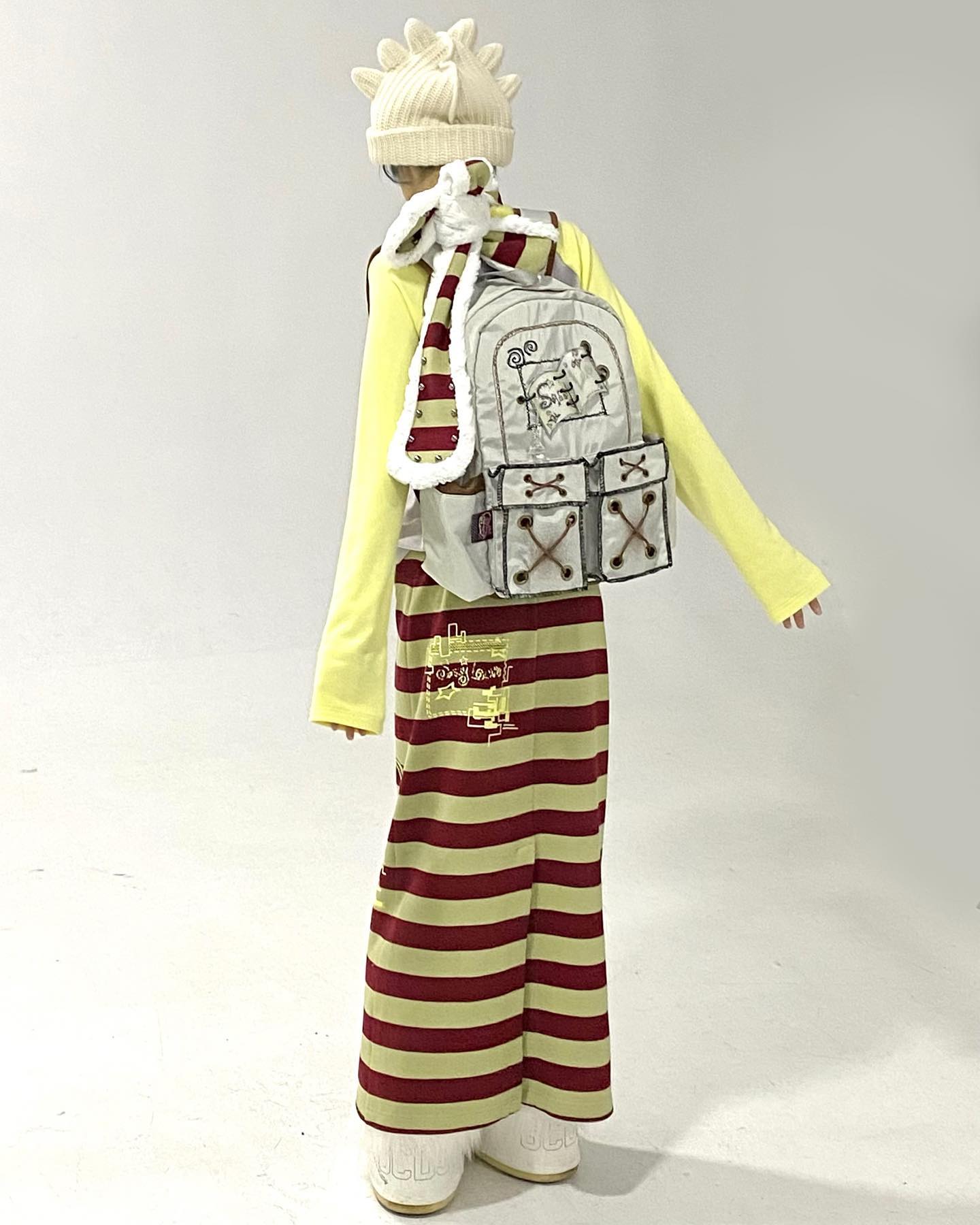 a full shot of a model wearing a unique outfit. the model is wearing a yellow longsleeve shirt, a long red and yellow striped skirt, a beige knitted hat with unique detailing, a fluffy scarf, and a gray backpack with illustrations on it. the model is wearing white shoes. the background is plain white. the style is unique, quirky, and artistic.