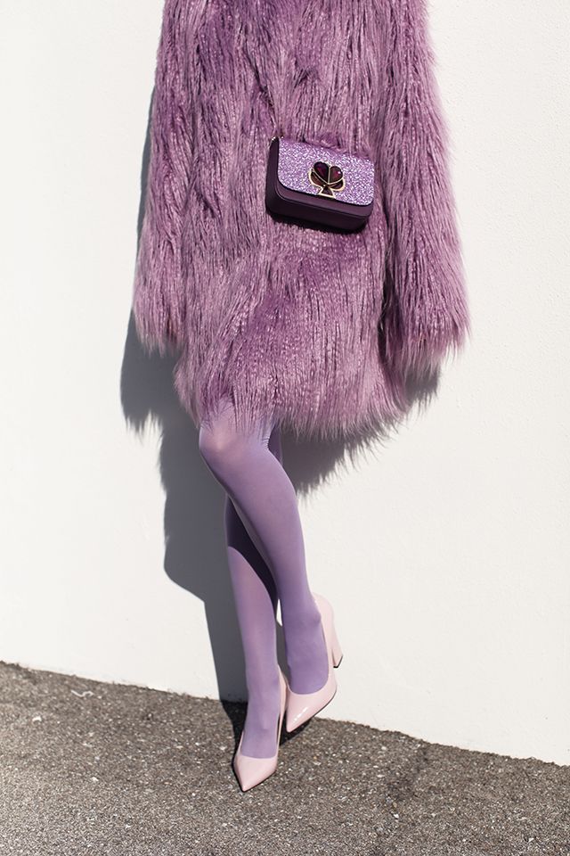 a woman is standing against a white wall wearing a lilac fluffy faux fur coat, matching lilac tights, and light pink heels. she is carrying a small purple and glitter kate spade purse. the overall look is a monochrome pastel purple outfit, stylish and coordinated.  the setting appears to be outdoors in sunlight.