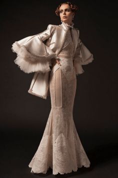 a woman in a white lace dress with a ruffled jacket poses for the camera. the dress is a vintage style with a long, flowing skirt and a fitted bodice. the woman is wearing her hair up and has a natural makeup look. the backdrop is a plain, dark background.