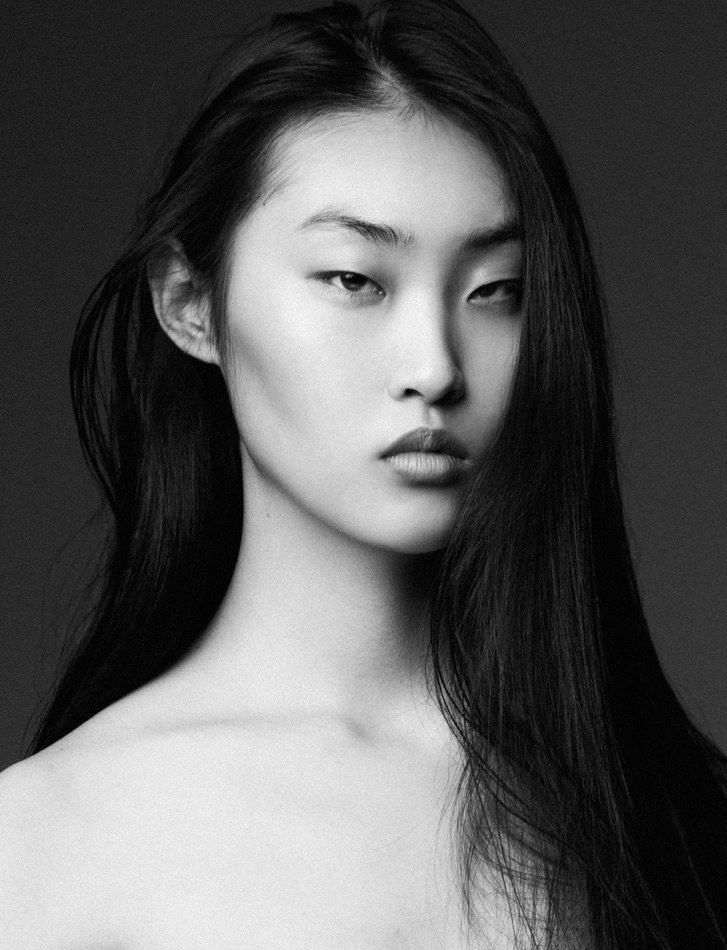 a striking black and white portrait of a young asian woman with long, straight hair. she has a serious expression on her face and is looking directly at the camera. the lighting is dramatic, highlighting her features. the background is a solid, dark gray.