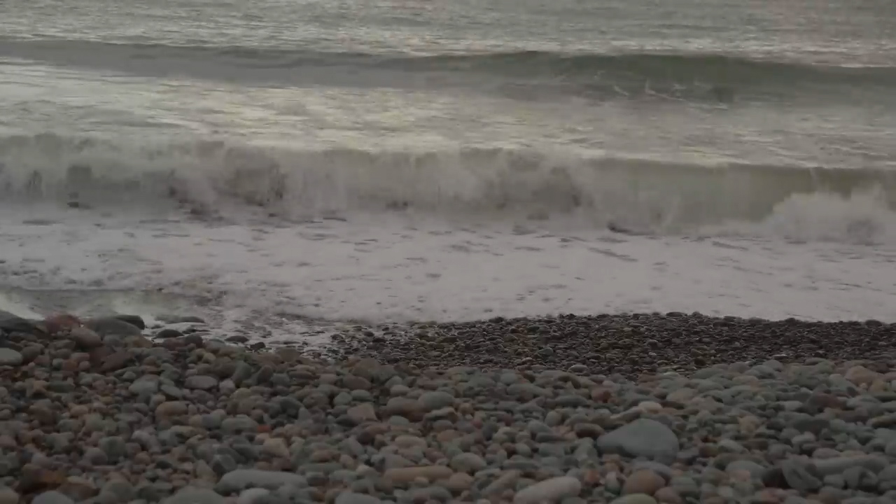the image shows a rocky beach with waves crashing on the shore. the beach is made up of small, smooth rocks in varying shades of gray. the waves are white and foamy, and the ocean in the background is a dark grayblue. the overall lighting is overcast and muted, creating a sense of tranquility and peace.