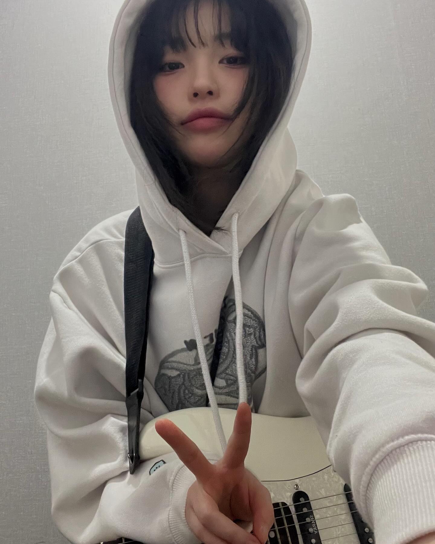 a young woman with dark hair and bangs poses for a selfie while holding a white electric guitar. she's wearing a white hoodie and gives a peace sign with her right hand. the image has a soft, naturally lit aesthetic and suggests an interest in music.