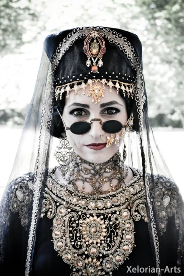 a woman in a black and white dress and veil, wearing ornate jewelry and sunglasses. she is looking at the camera with a mysterious expression. the image is in black and white and has a vintage feel.
