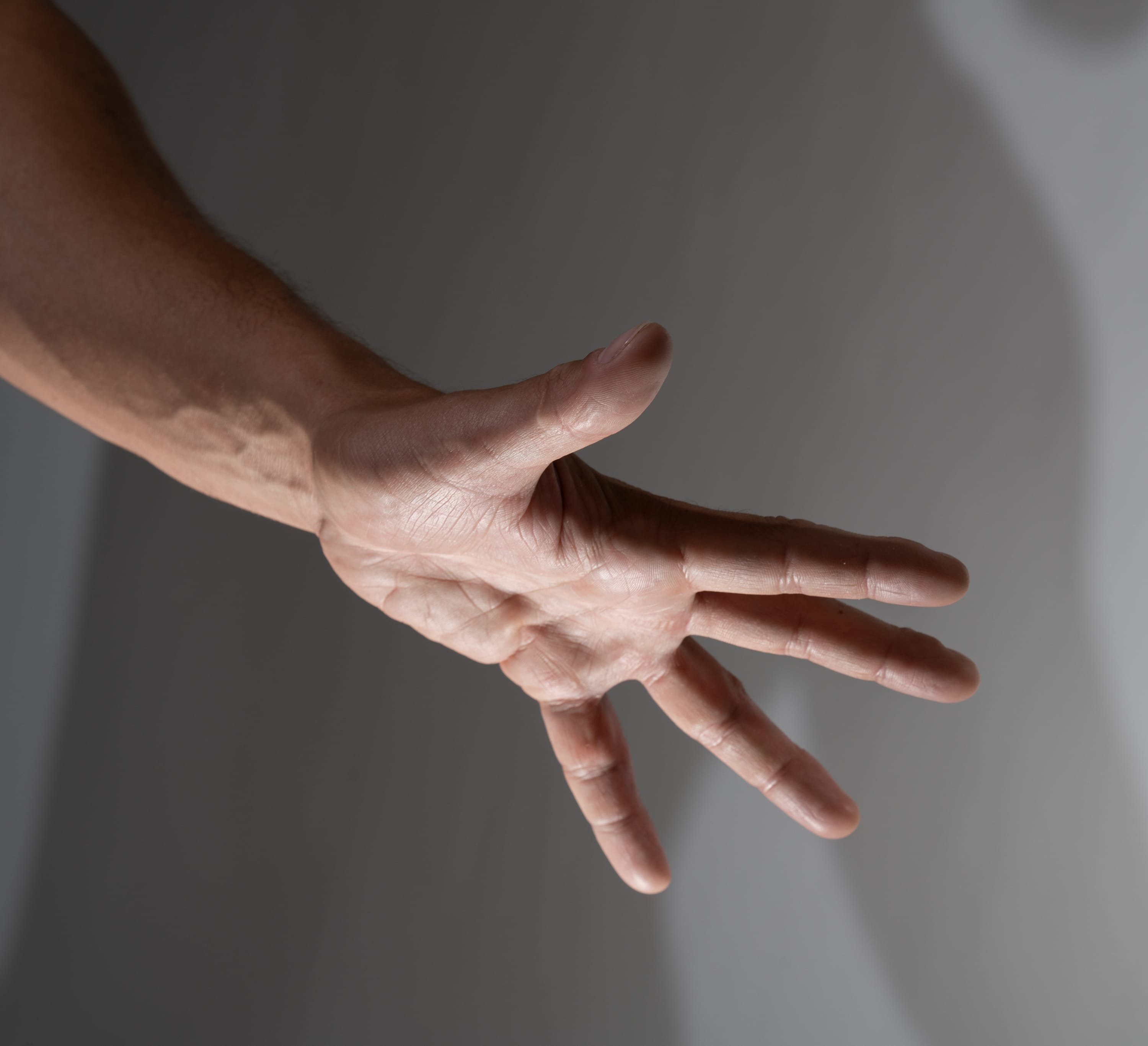 a closeup image of a man’s outstretched hand against a gray background. the hand is welllit, revealing details of the skin and veins. the fingers are slightly spread apart, suggesting a reaching or grasping motion.
