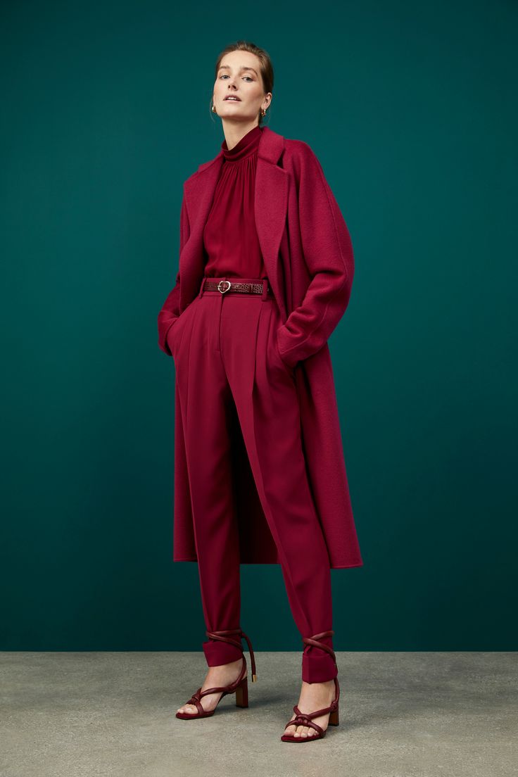 a fullbody shot of a woman model posing against a teal background. she is wearing a maroon turtleneck sweater, highwaisted pleated trousers, a long maroon coat, and maroon sandals with ties around the ankles. the overall look is elegant and sophisticated, suitable for autumn or winter. the color coordination is monochromatic, with various shades of maroon against the teal backdrop.