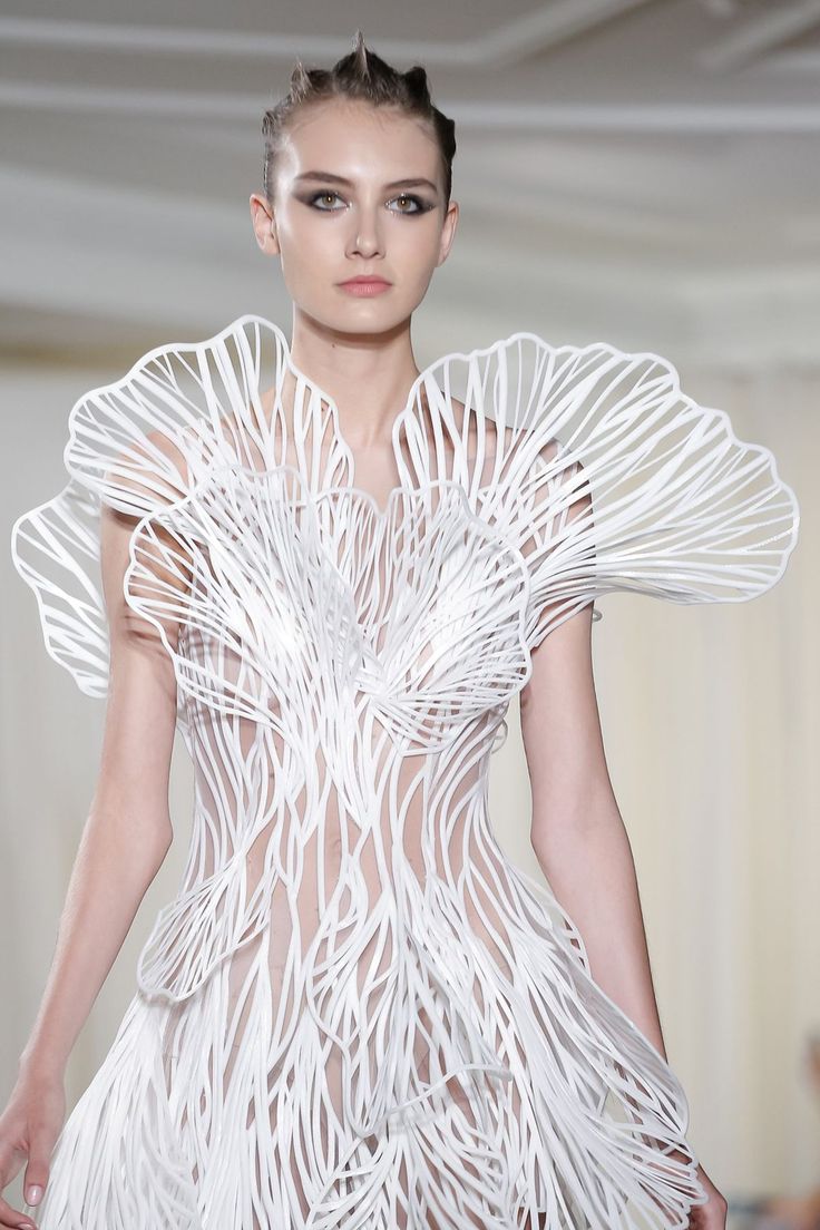 a woman in a white, intricate, 3d printed lace dress walks the runway. the dress is a futuristic and elegant piece, and it features a delicate, organic design.  the model is wearing her hair in a spiky style, adding to the avantgarde look.