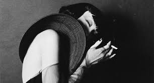 a black and white, closeup portrait of a woman wearing a large, widebrimmed hat. she is holding a cigarette in her hand, and her face is partially obscured by the hat and shadow. the overall mood is mysterious and sensual, evoking a classic film noir aesthetic. the image is sharply focused, with high contrast and deep blacks.  the woman appears stylish and elegant, with the clothing and pose suggesting a vintage or retro style, possibly from the 1940s or 1950s.