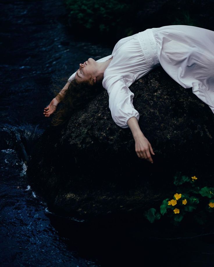 a woman in a white dress is laying on a dark rock in a river. she is facing upwards with her arms outstretched. the river flows around the rock, and there are green plants with small yellow flowers at the base of the rock. the background is a dark forest.