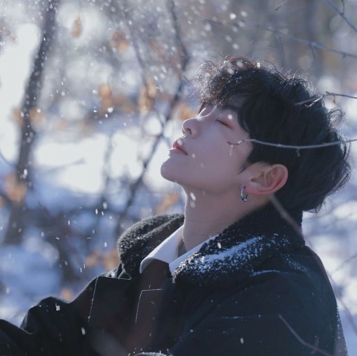 the image shows a young man, potentially a kpop idol,  in a winter setting. he is looking up, with his eyes closed, as snowflakes fall around him. he is wearing a black coat with a white collar.  the background is a blur of snowcovered trees and branches. the overall feel of the image is peaceful and serene. the soft, natural light contributes to the tranquil atmosphere.