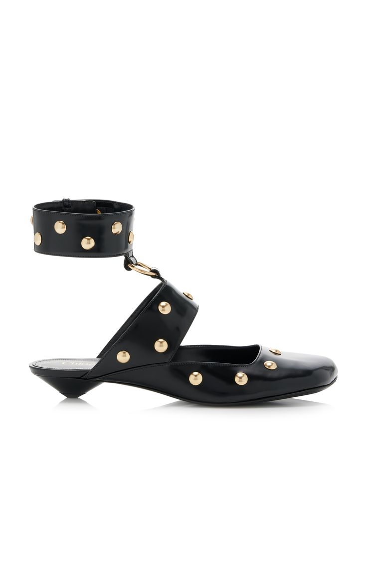 the image shows a pair of black leather mules with a distinctive design.  the shoes are adorned with goldtoned studs across the vamp and ankle strap.  they feature a low kitten heel and a pointed toe. a black leather ankle strap with a gold buckle secures the shoe around the ankle. the shoes are positioned on a white background.