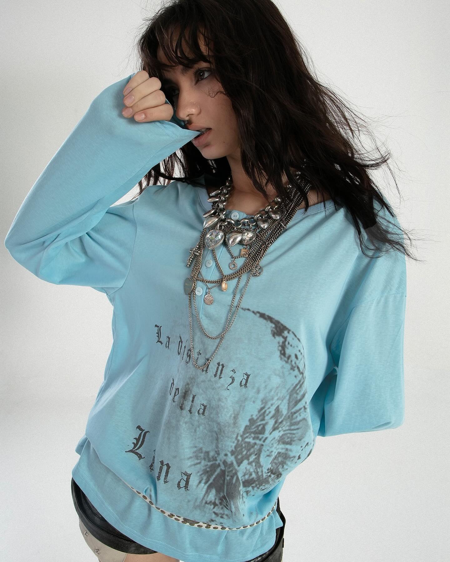 a young woman with long dark hair is modeling a light blue, oversized longsleeve shirt. the shirt features a graphic print of a moon with text in a gothicstyle font in italian, which translates to "the distance of the moon". she is wearing several layered silver necklaces. the overall style is grunge and vintageinspired. the image is shot against a plain white background.