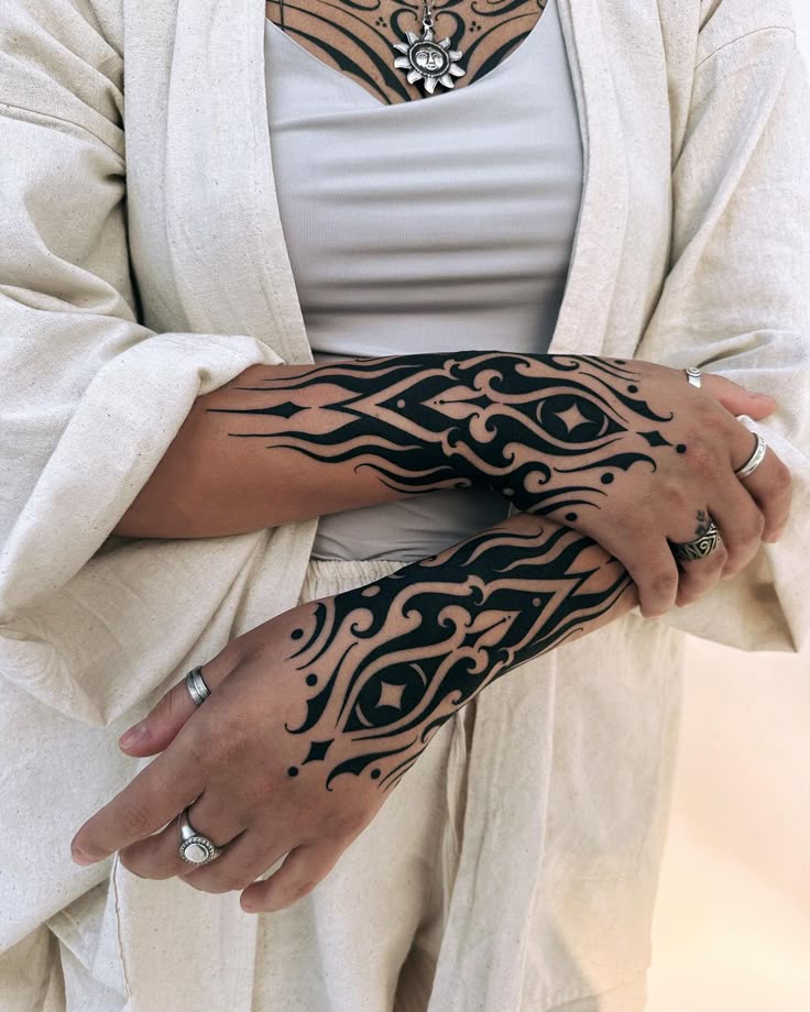 a woman with tribal tattoos on her arms and hands is seen wearing an offwhite shirt over a white tank top. she is wearing several rings on her fingers and a necklace. she also has a chest tattoo.