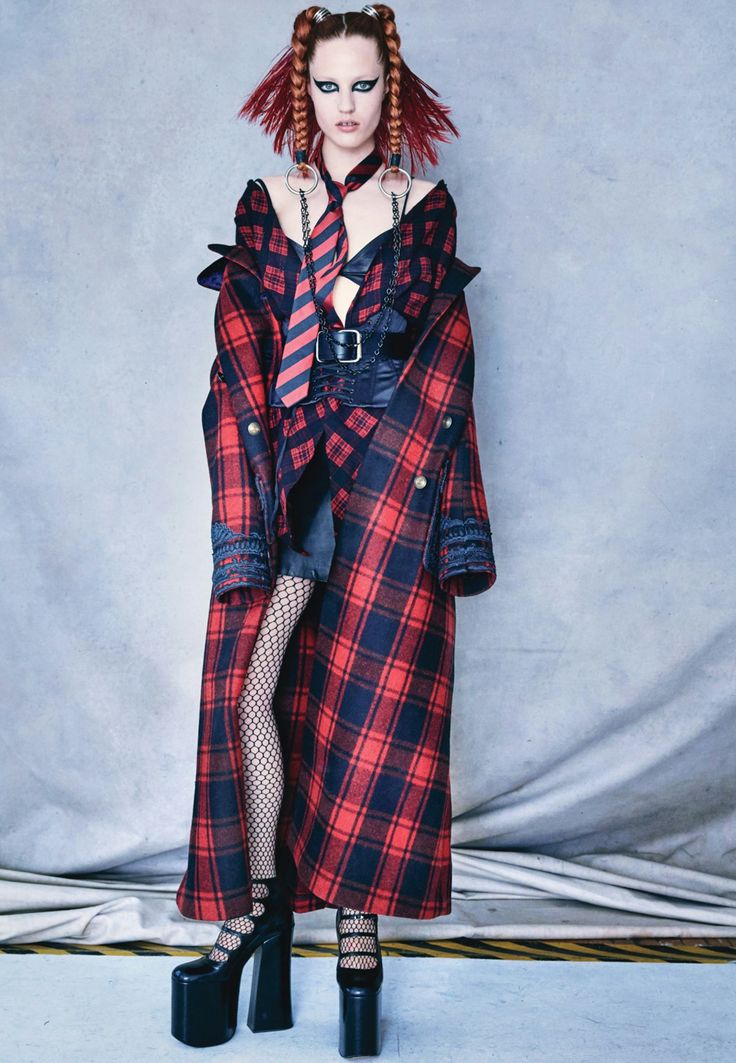 a woman models a red and black plaid coat with black platform shoes and fishnet stockings. she wears a tie and suspenders, and her hair is styled in braids with hoops. she has dramatic makeup, and a corset belt completes the look. the style is edgy and gothic.