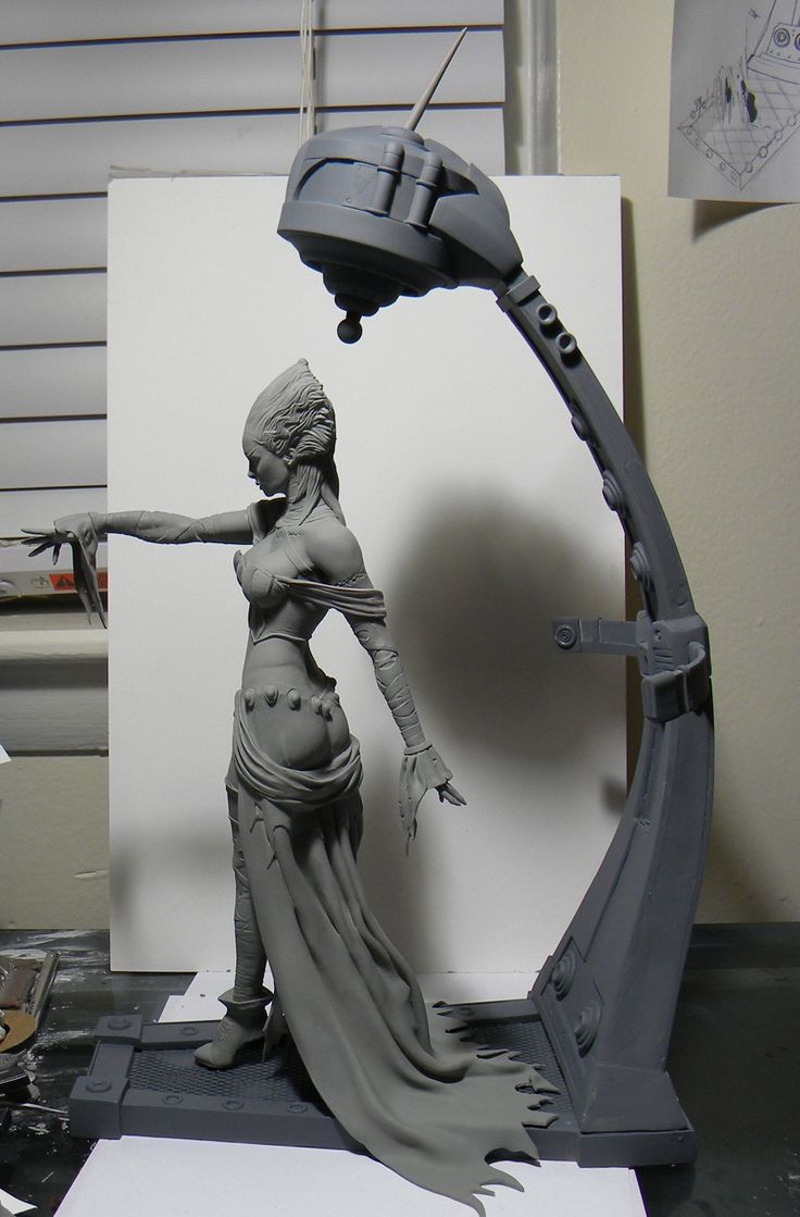 a grayscale unpainted sculpture of a woman with a futuristic headdress stands in front of a white board. the sculpture appears to be in progress, as it is surrounded by sculpting tools and materials. the woman is wearing a long, flowing gown and her arms are outstretched. the background is a white wall. the lighting is artificial and comes from the left side of the image.