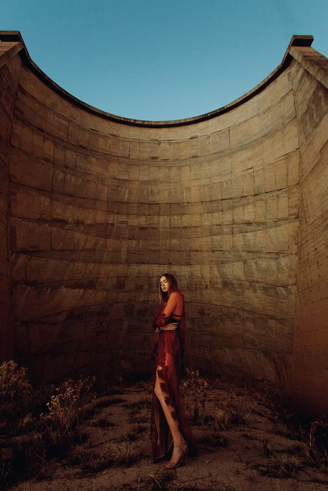 a woman in a flowing red dress stands in the center of a circular stone structure. the warm, natural light of the setting sun casts long shadows, highlighting the texture of the stone walls and the delicate fabric of her dress. the backdrop of a clear blue sky emphasizes the vastness of the desert environment. the woman's pose and attire suggest a bohemian fashion shoot, capturing a sense of freedom and natural beauty.