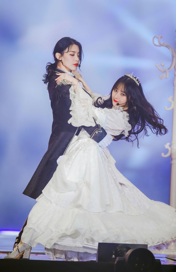 two women are on stage performing a dance routine. one woman is wearing a white dress with a long, flowing skirt. the other woman is wearing a black coat with a long, flowing skirt. they are both looking at the camera and smiling. the women are dancing gracefully and looking stylish. they seem to be having a lot of fun.