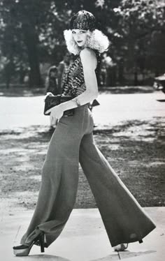 a woman in wideleg pants and a hat poses for a photo shoot. she is wearing sunglasses and holding a purse. the photograph is taken from the front and the woman is looking to the right. the photo is likely from the 1970s.