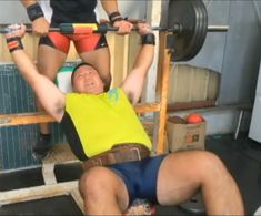 the image shows two men in a home gym environment.  one man, wearing a yellow shirt and blue shorts, is lying on a weight bench and lifting a barbell.  he is performing a bench press exercise.  the other man, wearing black and red shorts, is standing behind the bench, spotting the lifter. the lighting appears to be natural light coming from a door or window to the right, creating a bright but slightly shadowed scene. the perspective is at eyelevel and slightly offcenter, giving the viewer a good view of the action.