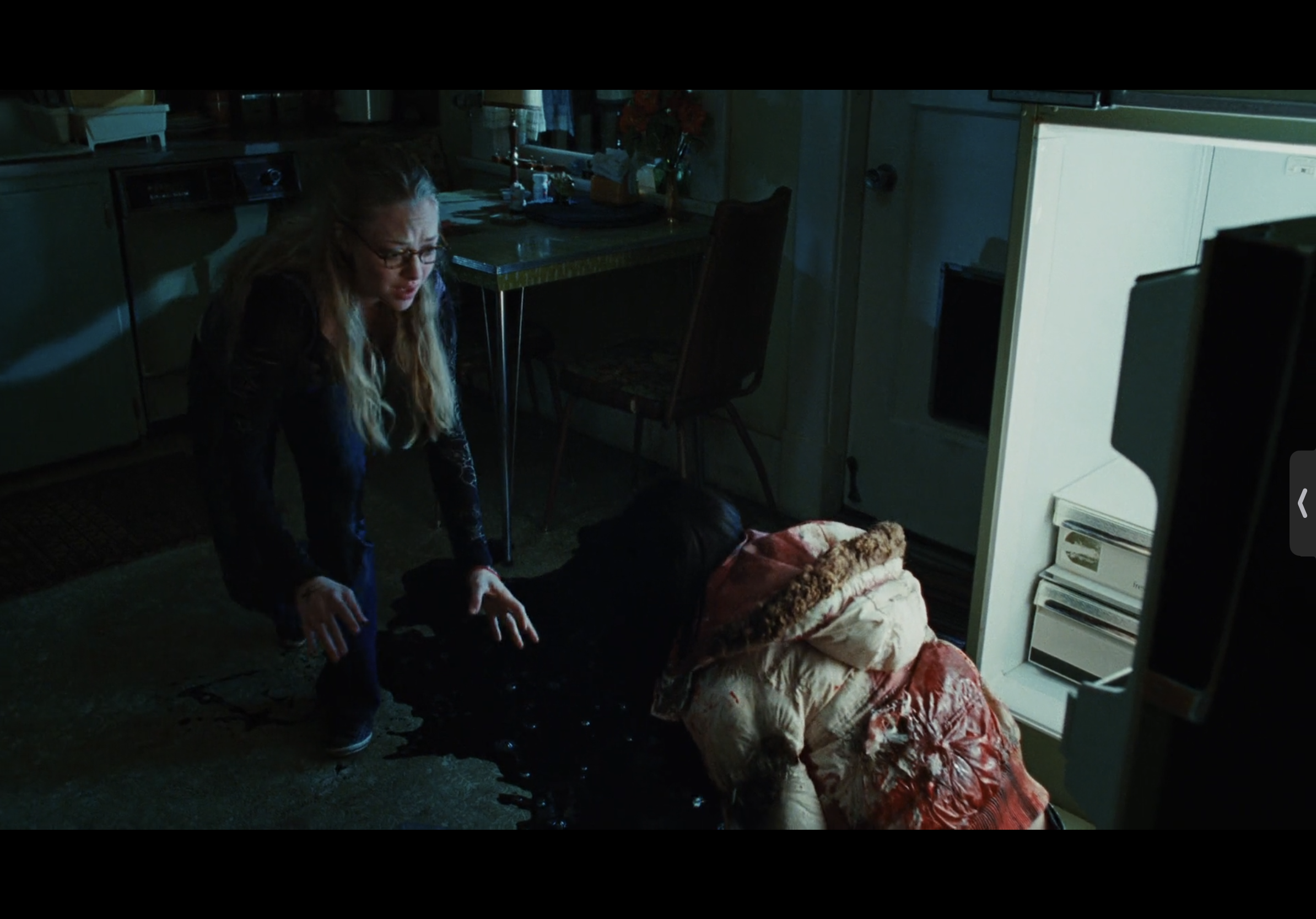 a woman in a black jacket and glasses stands over a dead body covered in blood on the floor of a kitchen. the woman looks shocked and scared.  there is a refrigerator on the right side of the image. the image is dark and shadowy, creating a sense of horror and dread.