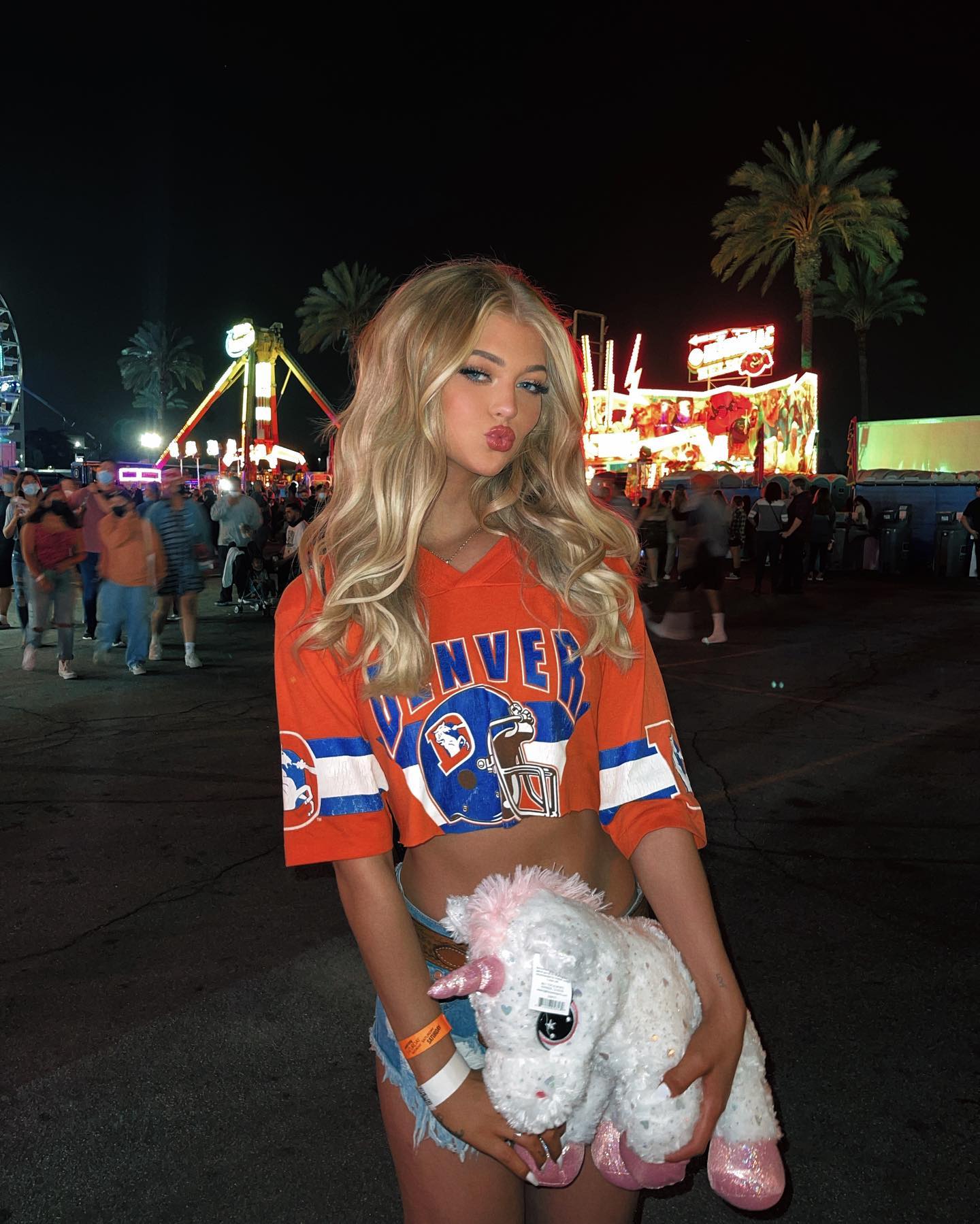 a young blonde woman stands at a fair, wearing a denver broncos shirt cropped at the midriff and denim shorts. she holds a white unicorn plushie. the background shows a busy fair with various rides, food stalls, and crowds of people under the night sky. palm trees are visible above the rides.