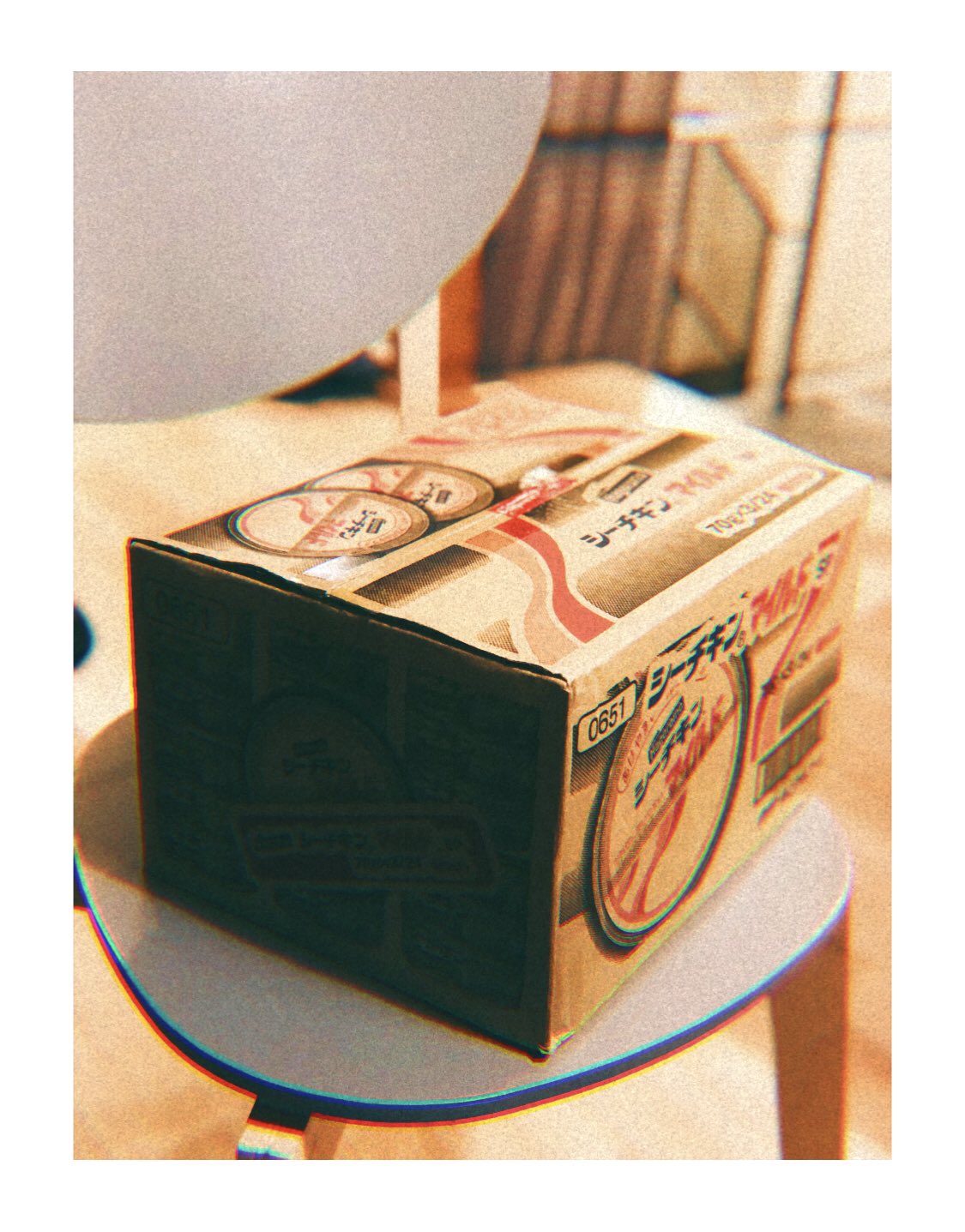 a vintage cardboard box with japanese writing sits on a white chair. the box appears to contain some sort of product, likely food, with a retro aesthetic. the image has a warm, nostalgic feel.