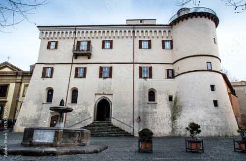 the image shows a grand, historic building with a tower, possibly a castle or a palatial residence, dominating the scene. the building has a lightcolored facade with multiple windows, some adorned with shutters, and a balcony. a rounded turret is attached to the side, adding to the building's architectural complexity. in front of the building is a stone fountain, and the surrounding area appears to be a town square or piazza, paved with stones. there are small plants in pots placed strategically around the square. the sky is overcast, and bare trees are visible in the upper corners of the image, suggesting it may be winter or early spring.
