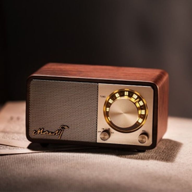 a vintagestyle wooden radio with a brown exterior, metal accents, and a round dial. the radio is sitting on a textured surface that appears to be a rug or fabric. it has a speaker grill on the front. the dial is illuminated, suggesting that it's turned on. the radio's design evokes a sense of nostalgia and classic style.