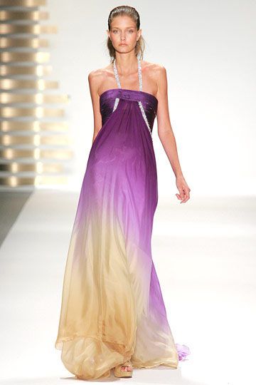 a model wearing a long ombre dress walks down a runway. the dress is a gradient of purple at the top to gold at the bottom. it is a halter top style and appears to be made of chiffon. the model has her hair pulled back in a ponytail.