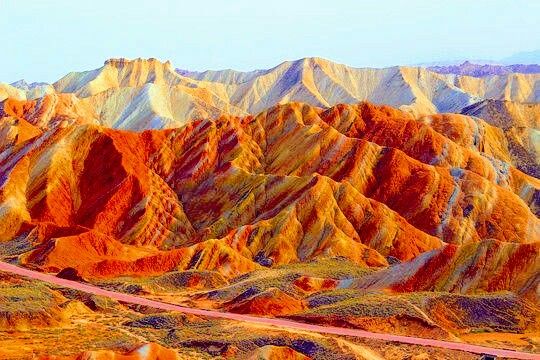 the image showcases the vibrant and colorful rock formations of the zhangye danxia landform geological park. the mountains display a stunning array of colors, including orange, red, yellow, and white, creating a unique and eyecatching landscape. a road cuts through the desert terrain, adding a sense of scale to the vast geological park. the sky is clear, emphasizing the natural beauty of the colorful hills.
