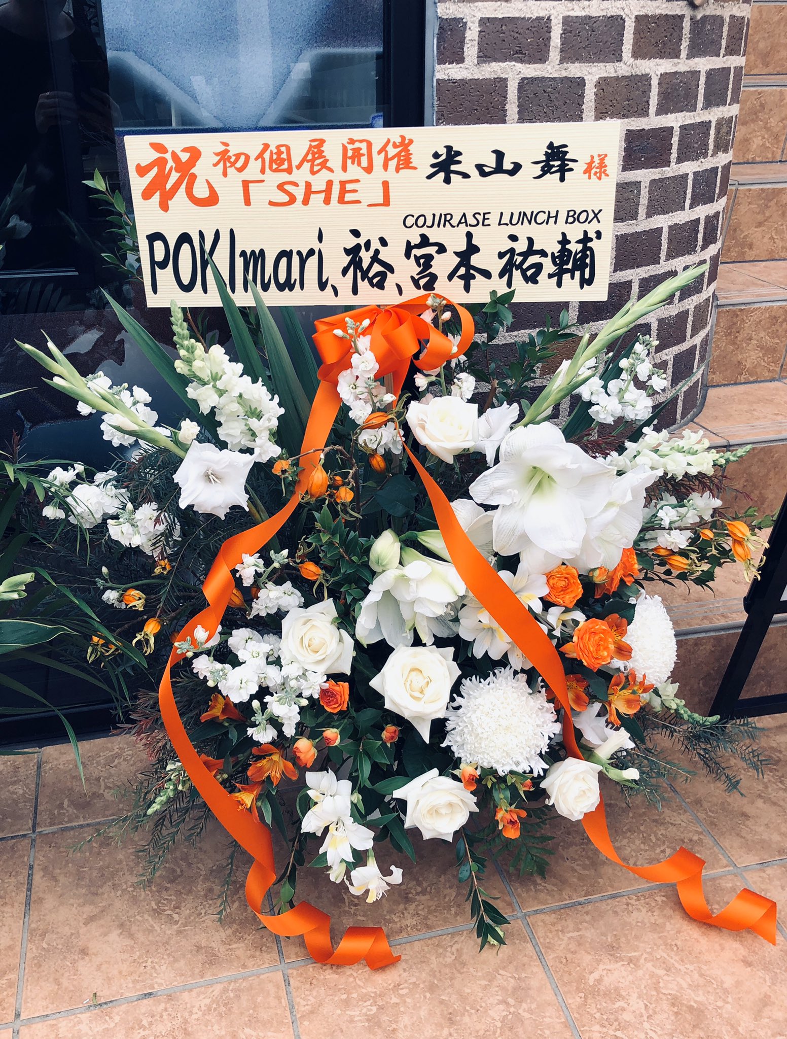 a cheerful display of orange and white flowers in front of a brick storefront, celebrating a grand opening.  a handwritten sign on an orange board congratulates someone on their first solo exhibition. the text is in japanese.