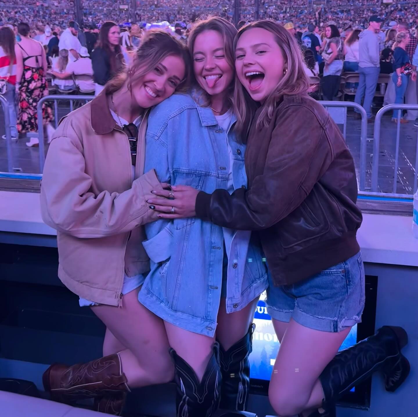 the image shows three young women, closely embracing each other at what appears to be a concert in a large stadium. the girls are all smiling and happy. one girl is wearing a light tan jacket, another a light blue denim jacket, and the third a brown leather jacket. two of the girls are wearing cowboy boots. the background is blurry but shows a large crowd of people at a concert venue. the focus is clearly on the three girls in the foreground. the overall atmosphere is joyful and celebratory.