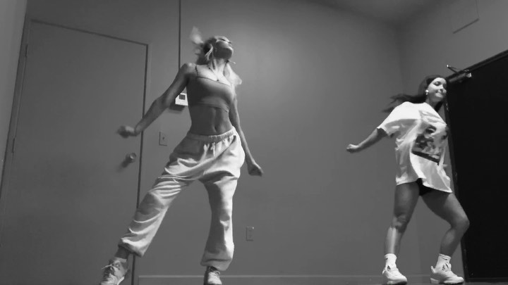two women are indoors, dancing. they are in black and white. one woman wears a top and loose pants with shoes. the other woman wears a tshirt with shorts and shoes with socks.