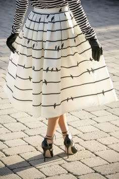 a woman wearing a white and black striped skirt with black heels stands on a cobblestone street. she is wearing black gloves and a white striped shirt. she is looking down at the ground.  the skirt is pleated and the heels are closed toe.