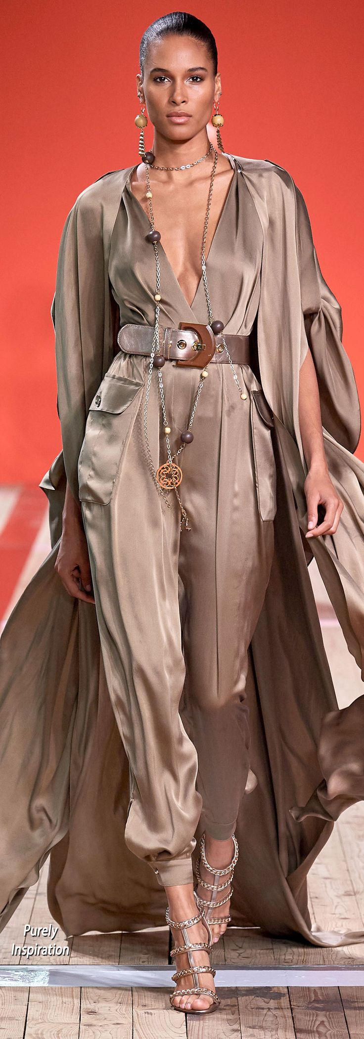a full shot of a female model on a runway. she is wearing a long, flowing beige jumpsuit with a matching long overcoat. she has on gold jewelry including long earrings, necklaces, and a belt.  her shoes are strappy high heels with embellishments.  the background is a solid orangered color.