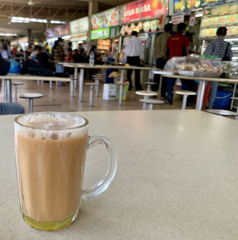 a closeup shot of a mug of teh tarik, a popular malaysian milk tea, sits on a table in a bustling hawker center or food court. the background is slightly out of focus but shows a busy scene with numerous food stalls, people eating, and a general atmosphere of a lively public eating area. the warm colors of the tea and the overall setting contribute to a sense of everyday life and local culture.