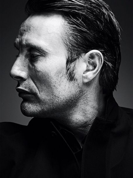 a black and white portrait of mads mikkelsen in profile, facing left. he has dark hair, stubble, and is wearing a suit. his expression is serious and intense. the image is tightly cropped around his head and shoulders.