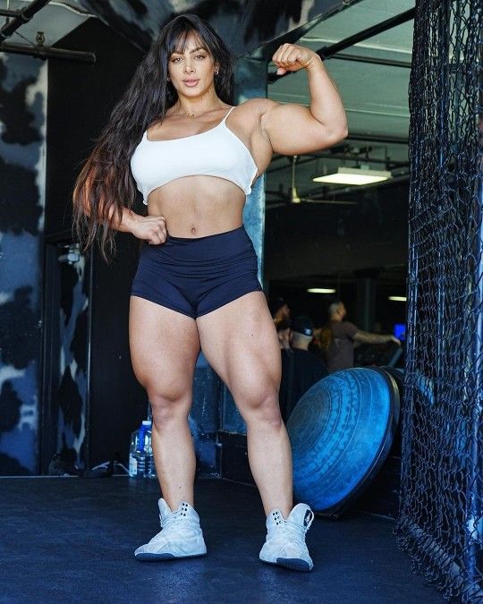 the image shows a muscular female bodybuilder posing in a gym. she's wearing a white sports bra and black shorts, showcasing her welldeveloped biceps, triceps, and especially her powerful legs and thighs. her long dark hair is loose, and she's wearing gray athletic sneakers. the background features gym equipment, including a bosu ball and a section of netting. the overall impression is one of strength, fitness, and athleticism.