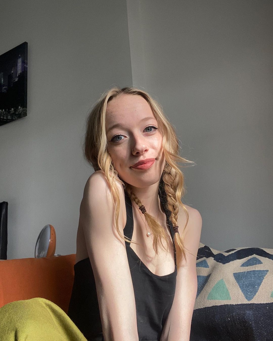 a young blonde woman with her hair in two braids is sitting on a couch, smiling at the camera. she's wearing a black tank top and green pants.  sunlight is illuminating her from the side. the background shows a simple home interior with a wall and a piece of artwork.