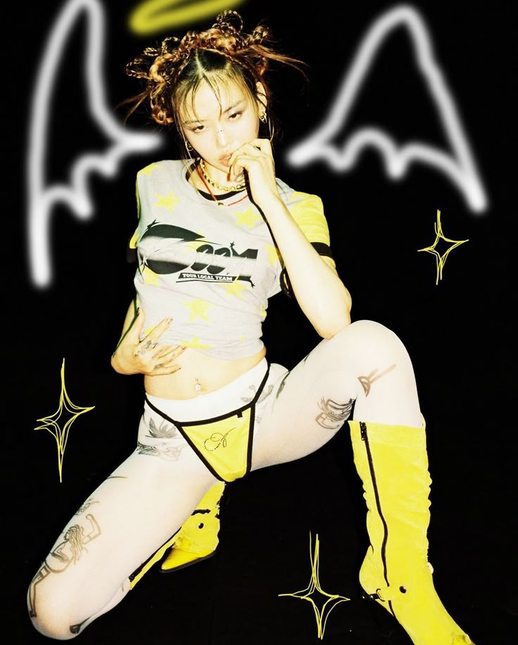 a woman in a gray tshirt, yellow lingerie, patterned tights, and yellow boots is posing against a black background with angel wings, a halo, and star graphics. she has multiple tattoos on her body and her hair is styled in buns. she is kneeling with one knee up and her hand on her stomach. she is looking down.