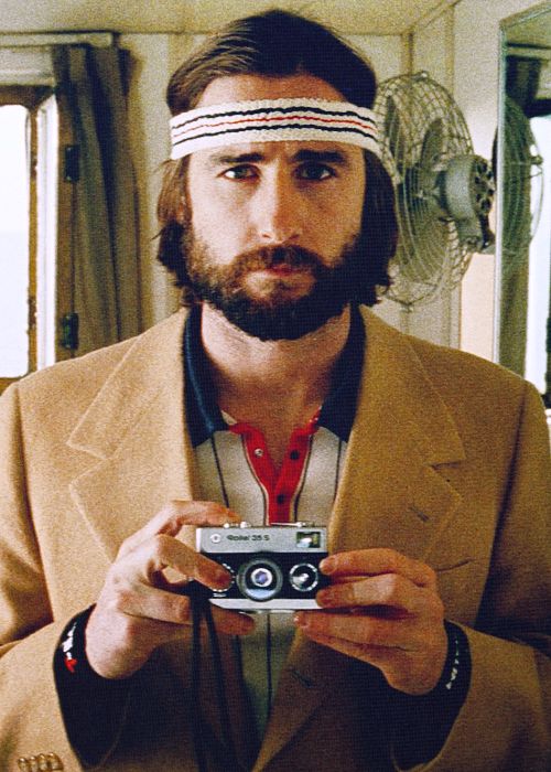 a man in a seventiesstyle coat, with a headband, mustache, and beard, holds a vintage film camera in his hands. he looks at the camera with a serious expression.  the background includes a wall, a window, and a fan. the man appears to be a photographer.