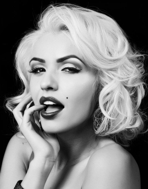 a black and white portrait of a glamorous blonde woman in a classic pinup style. she is wearing a simple black dress and has a stylish haircut. her hair is styled in loose curls, her makeup is perfect, and her expression is sultry and confident. her fingers are touching her lips and her eyes are looking directly at the viewer.