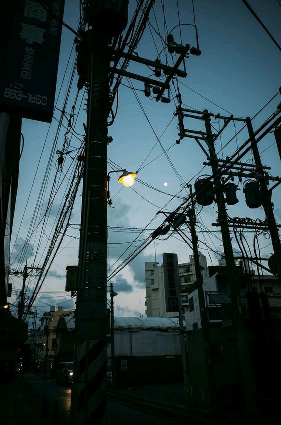 the image captures the ambiance of a japanese city street at dusk. the scene is dominated by a web of electrical wires strung between utility poles, creating a sense of urban density. a lone streetlamp casts a warm, yellow glow against the darkening sky, while the buildings and surrounding details fade into silhouette. the overall mood is atmospheric and cinematic, evoking a sense of mystery and tranquility.