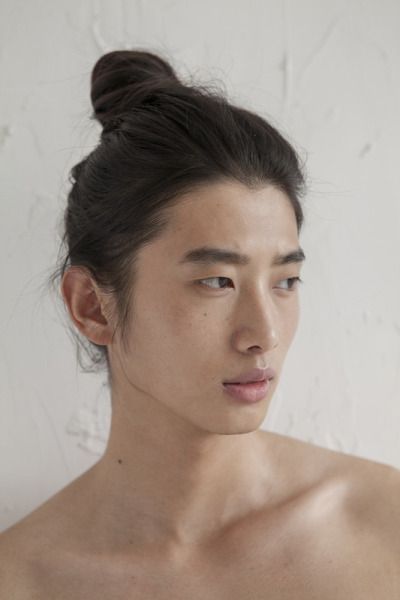 a closeup portrait of a young asian man with his dark hair styled in a high bun. he has bare shoulders and is set against a plain, neutralcolored background. his features are soft and appear androgynous.
